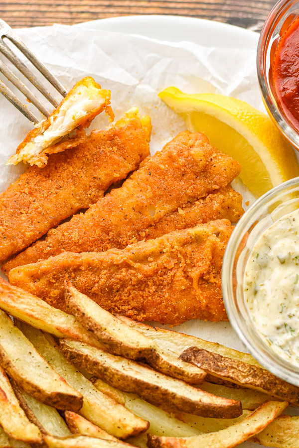 a fork with a piece of a fish fillet next to other breaded fillets, fries, and condiments 