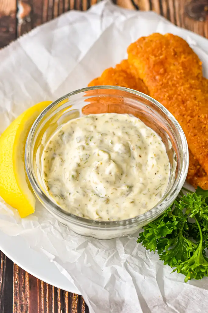 a bowl of low fodmap tartar sauce on white parchment paper with breaded fish, parsley, and a slice of lemon