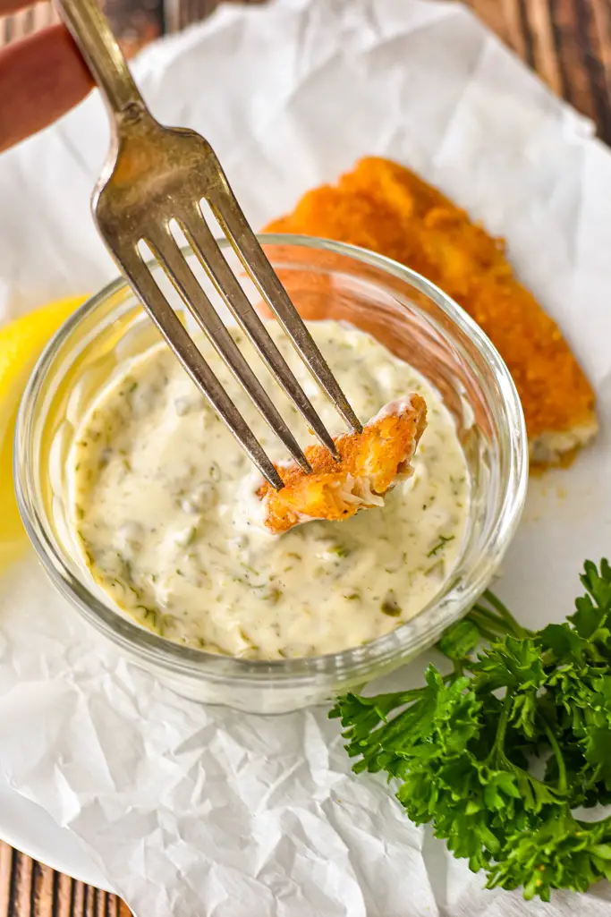 a fork dipping a piece of breaded fish into a small bowl of tartar sauce