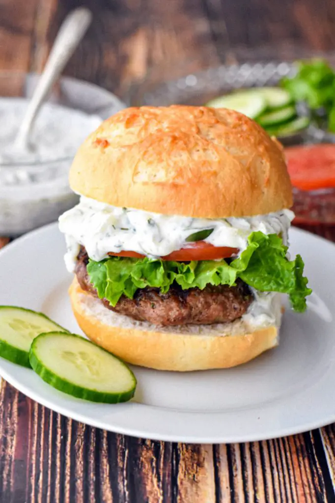 a low fodmap gyro burger topped with tzatziki and raw veggies on a gluten-free hamburger bun next to sliced cucumber on a white plate